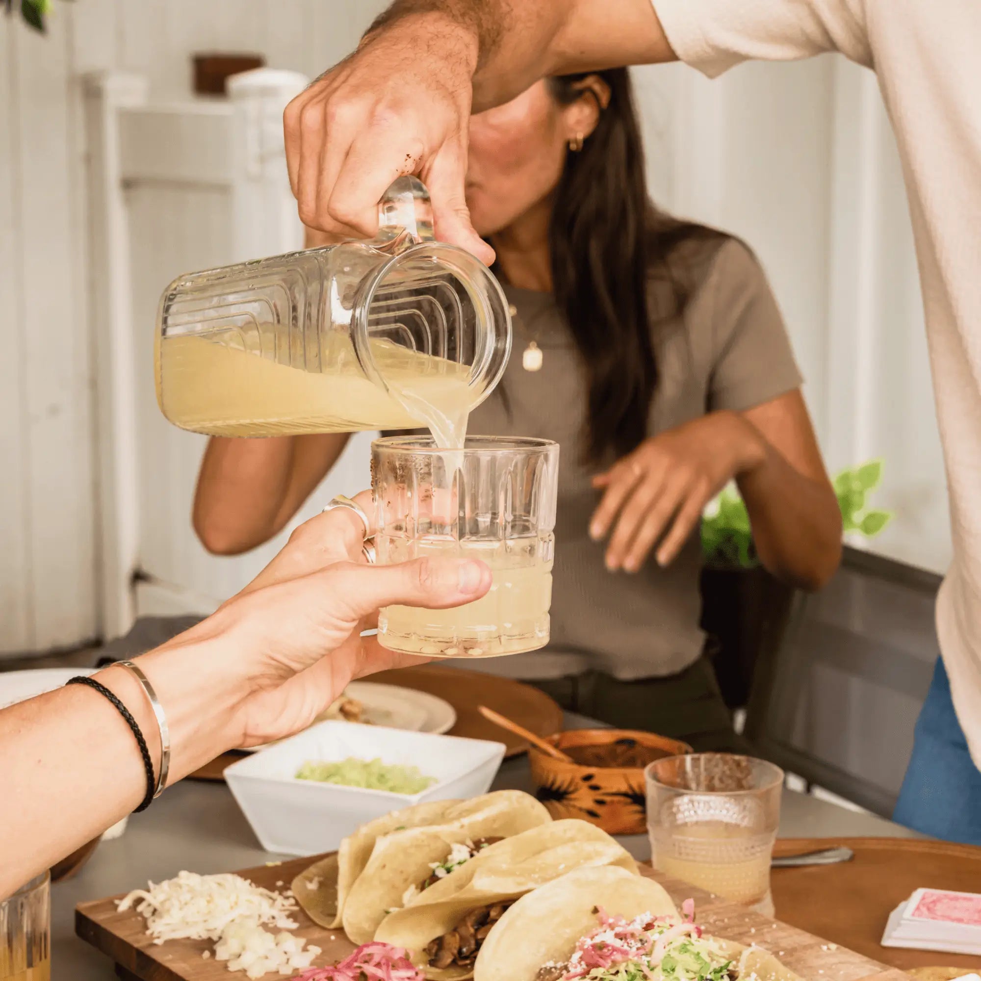 A group of friends enjoying drinks while a man pours classic margarita from a pitcher, with tacos and other food on the table